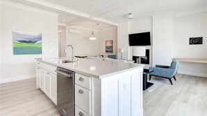 Una cucina moderna con mobili bianchi, lavastoviglie integrata e una grande isola. C'è un'area pranzo con una sedia blu e un televisore a schermo piatto montato a parete.