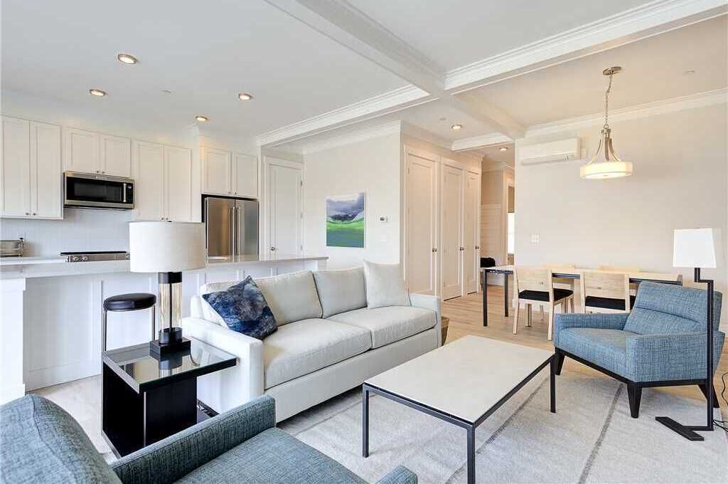 Ein modernes Wohnzimmer mit einer Couch, einem Couchtisch und Sesseln. Es gibt eine Küchenzeile mit einer Mikrowelle und einem Kühlschrank.