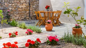 Een terras buiten met een stenen muur, een houten tafel en stoelen, een grote parasol en potplanten.