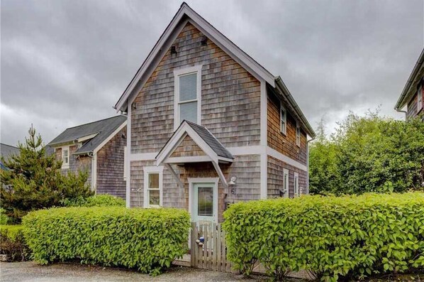 A two-story wooden house with a gabled roof, a front porch, and a small front yard with hedges.