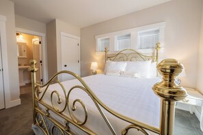 Une chambre à coucher avec un lit en métal doré, du linge de lit blanc, une table de chevet avec une lampe et une fenêtre avec des stores.