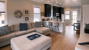 Una sala de estar moderna con un sofá seccional color beige, una mesita de centro y una cocina con armarios oscuros y una isla blanca.
