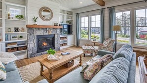 A cozy living room with a stone fireplace, wooden beams, and a large window offering a view of the outdoors. - Seabrook Sunset Retreat | Pet Friendly, Ocean View | 6BR, Sleeps 15, Sauna (Pacific Beach)