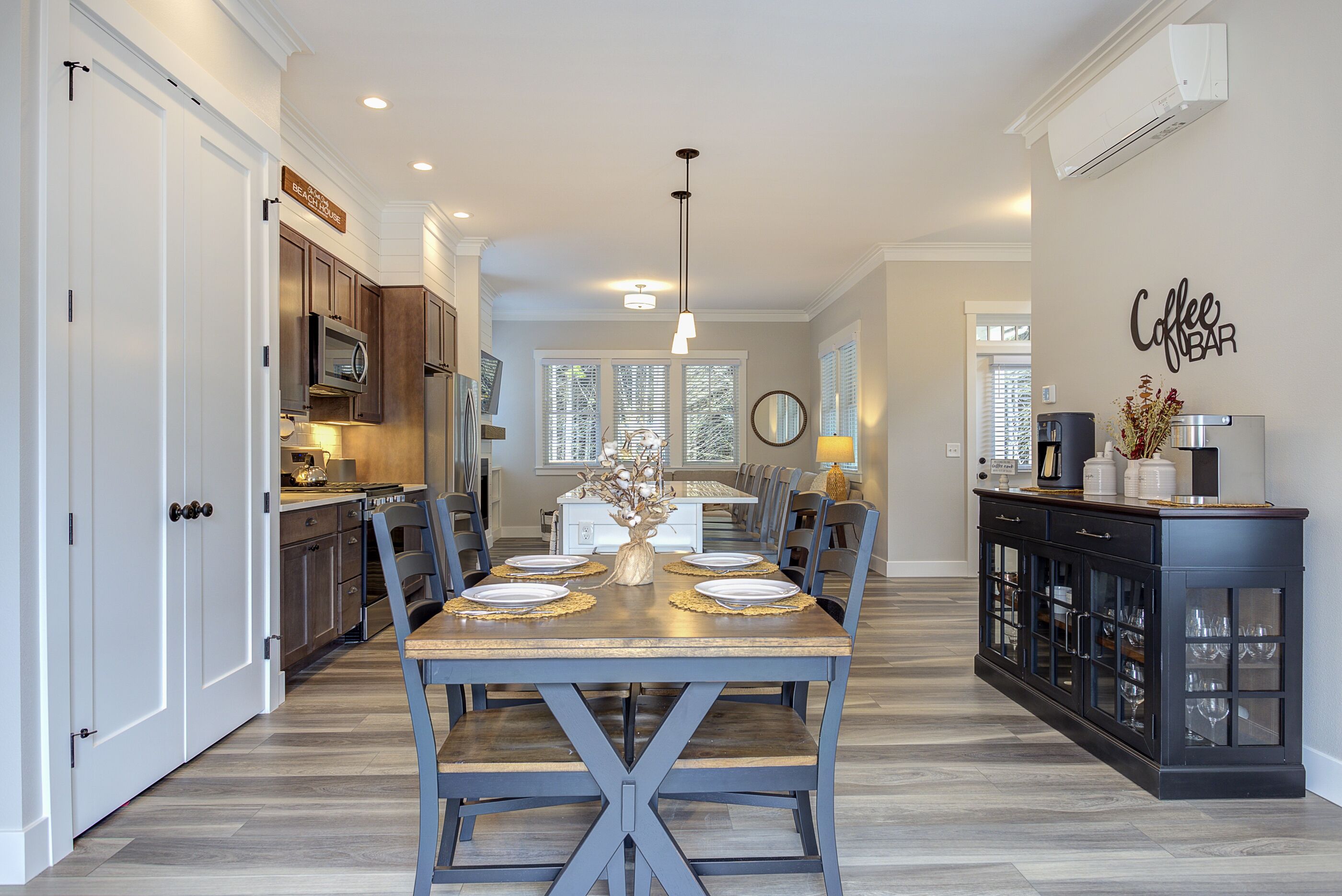 A dining area with a wooden table set for six, flanked by dark blue chairs, a kitchen with stainless steel appliances, and a coffee bar with a sign.