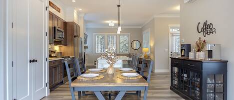 A dining area with a wooden table set for six, flanked by dark blue chairs, a kitchen with stainless steel appliances, and a coffee bar with a sign.