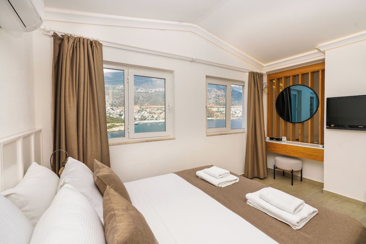 A hotel room with a bed, two windows with curtains, a desk with a chair, a flat-screen TV, and a view of a town and mountains.