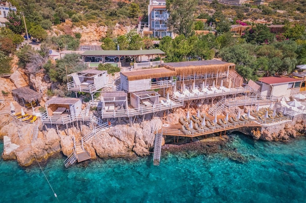 An aerial view of a coastal resort with multiple overwater bungalows, clear turquoise waters, and rocky terrain.