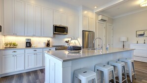 A modern kitchen with white cabinetry, a stainless steel refrigerator, a microwave, and a large island with bar stools. - Seabrook The Penthouse | Ocean View, Pet Friendly | 1BR, Sleeps 4, Rooftop BBQ (Pacific Beach)