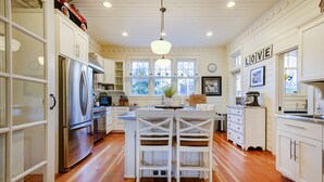 A kitchen with white cabinets, a stainless steel refrigerator, wooden flooring, and a central island with two chairs. - Seabrook Washington House | Piano, Sleeps 16+, 7BR | Historic Design, Crescent P (Pacific Beach)