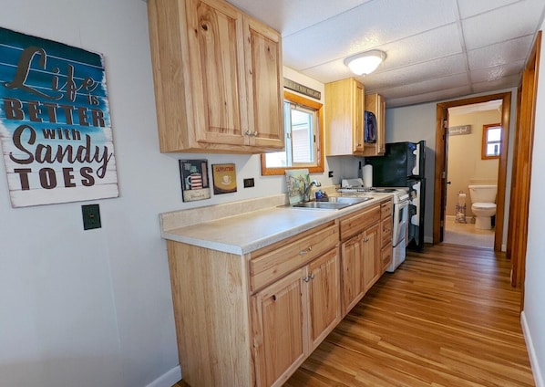 A kitchen with wooden cabinets, a countertop, a sink, a stove, and a refrigerator. There is a window above the sink and a door leading to another room.