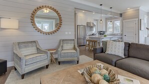 Una sala de estar moderna con dos sillones, un sofá, una mesa de centro con objetos decorativos, un espejo grande con marco de madera y una cocina al fondo.