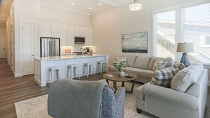 A modern kitchen with white cabinets, stainless steel appliances, and a central island with stools. There is a living area with a sofa, a coffee table, and a painting on the wall. - Seabrook Windjammer | Ocean View, Rooftop Deck 2BR | Sleeps 4, Town Center (Pacific Beach)