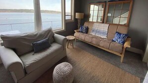 Ein gemütliches Wohnzimmer mit einem beigen Sofa, einem kleinen Sessel und einem runden Hocker. Große Fenster bieten Blick auf das Wasser.
