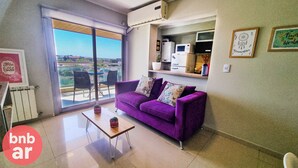 A modern living room with a purple sofa, a small table, and a large window offering a city view. - Beautiful condo with Swimming Pool  and 24/7 security (Córdoba)