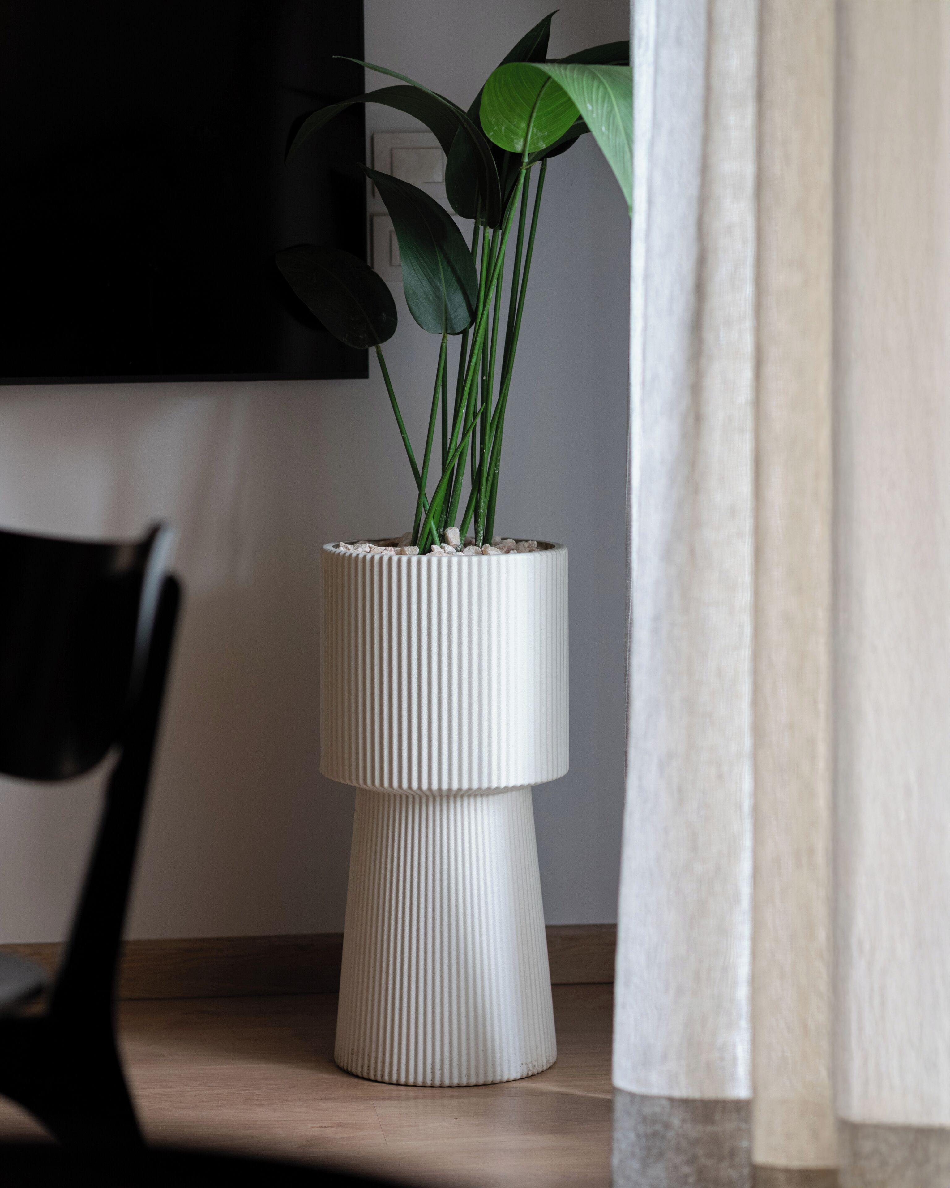 A white ribbed planter with green plants on a wooden floor, next to a black chair and a white wall.