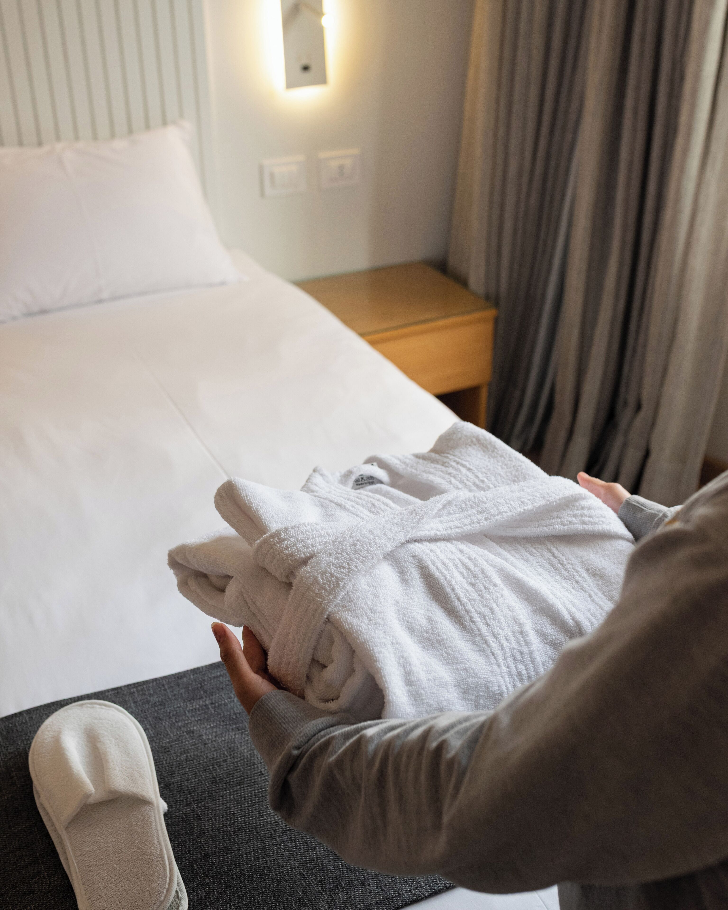 Un lit bien fait, avec du linge de lit blanc, une table de chevet en bois sur laquelle se trouve une lampe, et une paire de chaussons posés sur le sol.