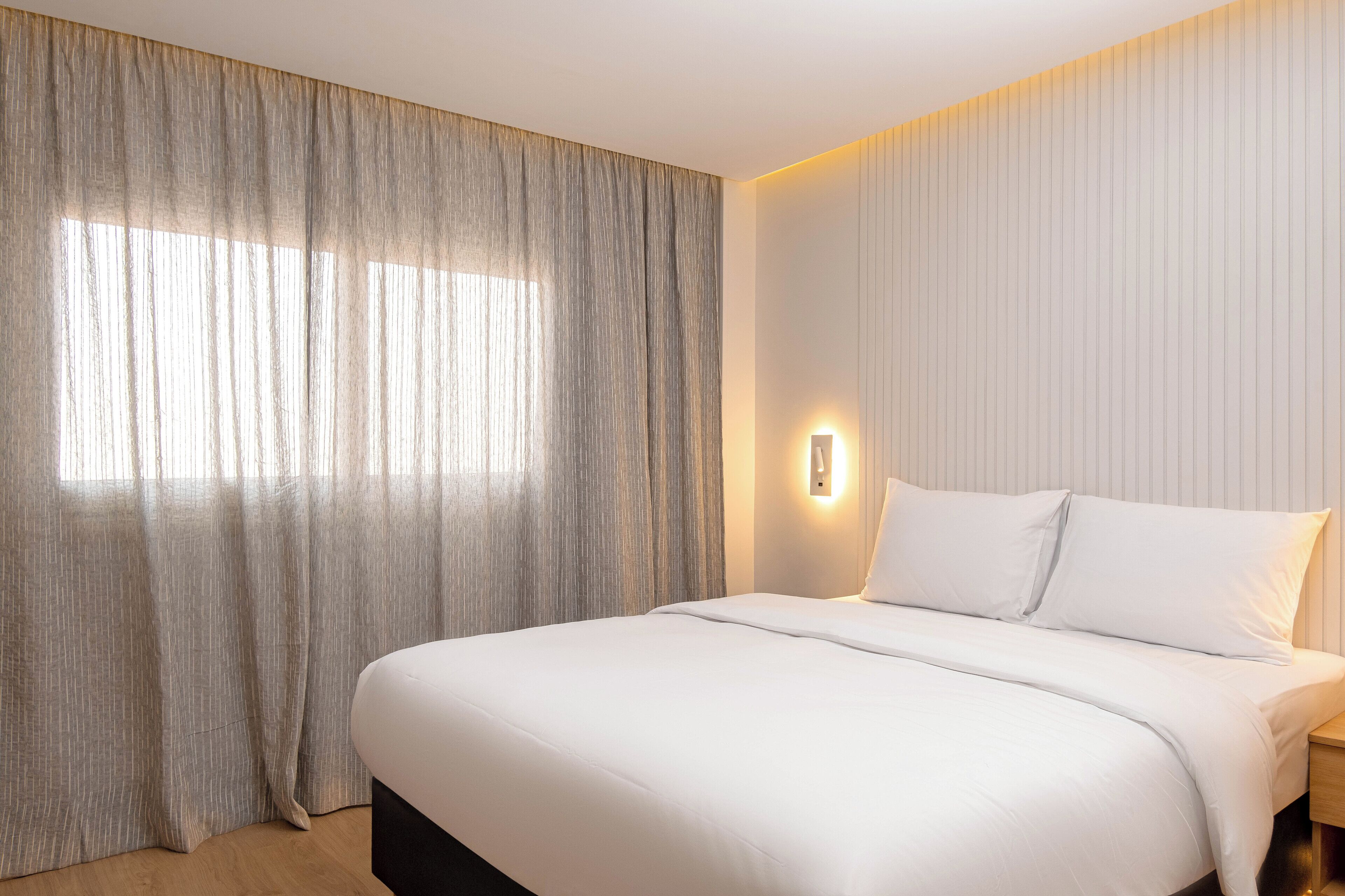 Une chambre d’hôtel avec un grand lit, une table de chevet, une lampe fixée au mur et une fenêtre avec des rideaux légers.