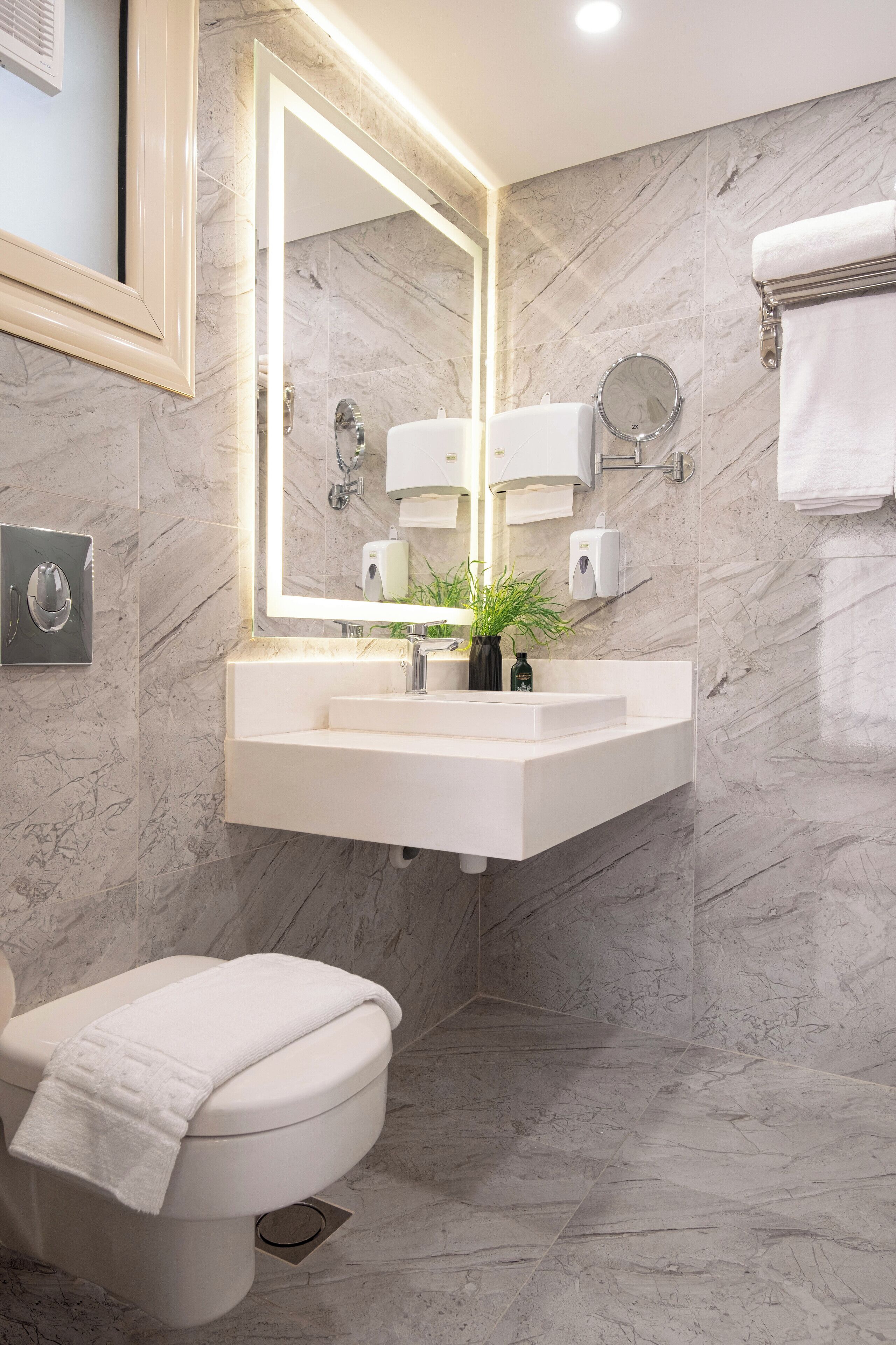 A modern bathroom with a marble floor, a white sink with a mirror above it, a toilet with a towel on it, and a wall-mounted soap dispenser.