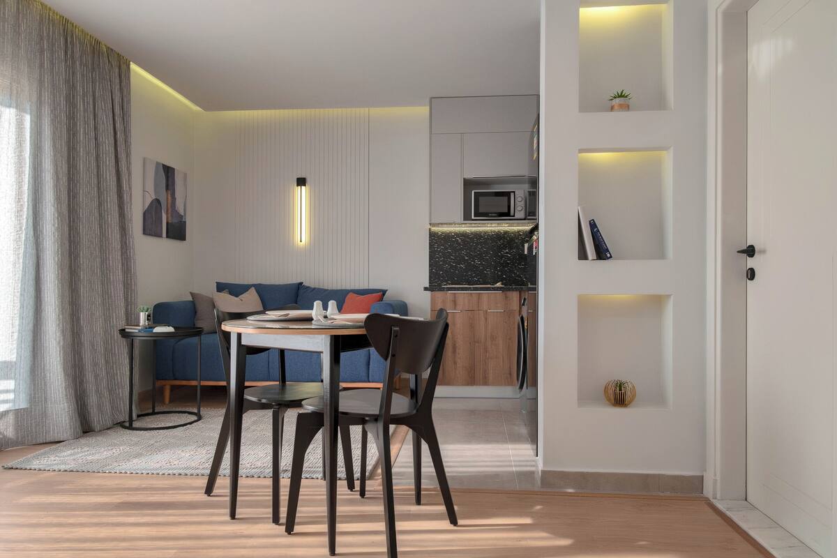 A modern living room with a dining table, chairs, a sofa, and a kitchen area with built-in appliances.