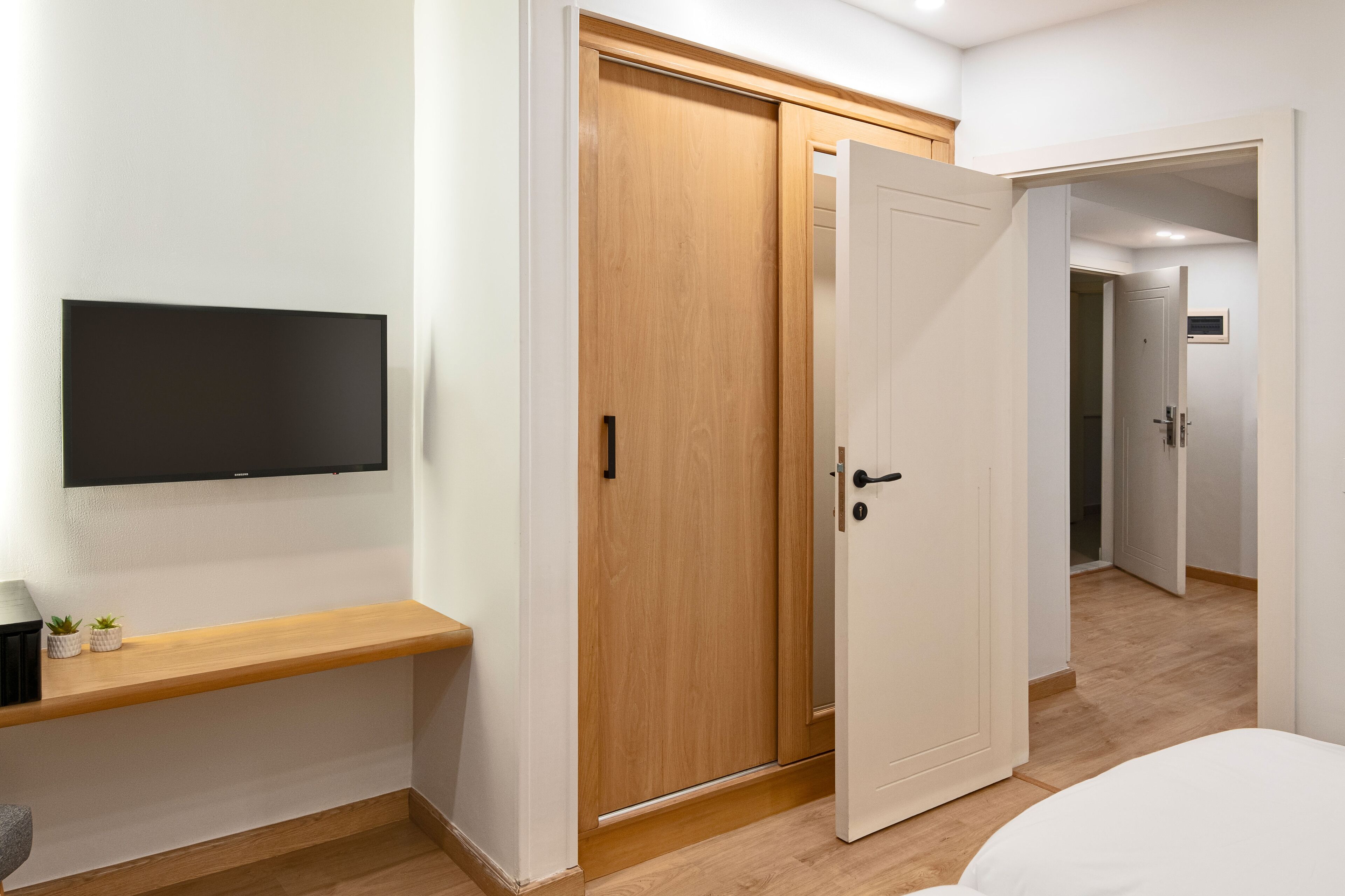 A bedroom with a bed, a TV mounted on the wall, a wooden wardrobe, and a door leading to another room.