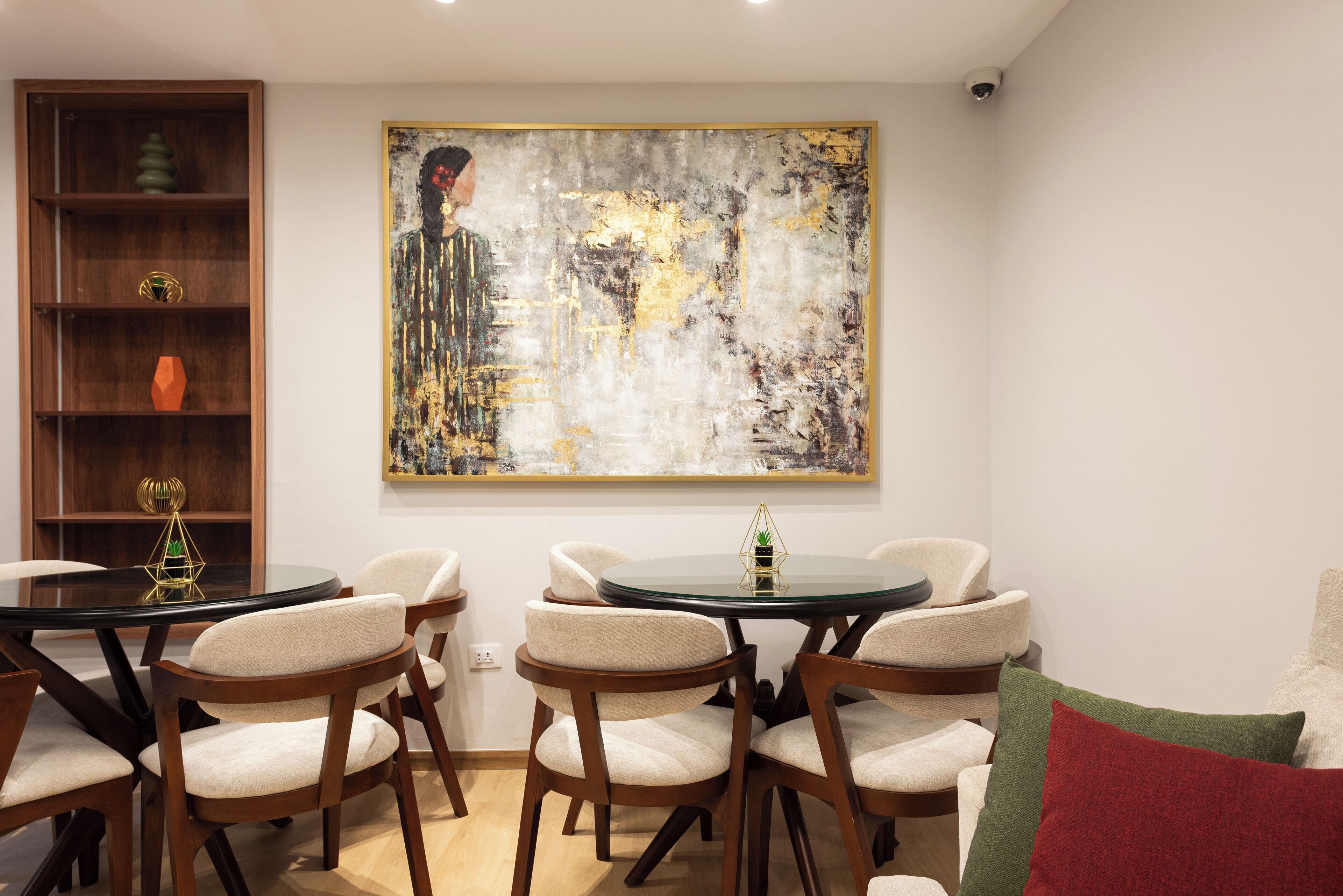Een eetgedeelte met een ronde glazen tafel, zes gestoffeerde stoelen, een boekenkast met decoratieve objecten, en een groot abstract schilderij aan de muur.