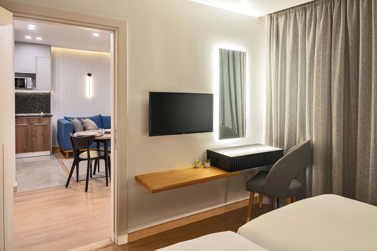 Une chambre d’hôtel moderne avec un téléviseur à écran plat fixé au mur, une étagère en bois ornée d’une petite plante, un fauteuil gris et un lit.