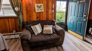 A cozy room with a brown leather sofa, a wooden wall, a framed butterfly artwork, a door with a 'No Smoking' sign, and a window with curtains. - Riverside Motel (Lewiston)