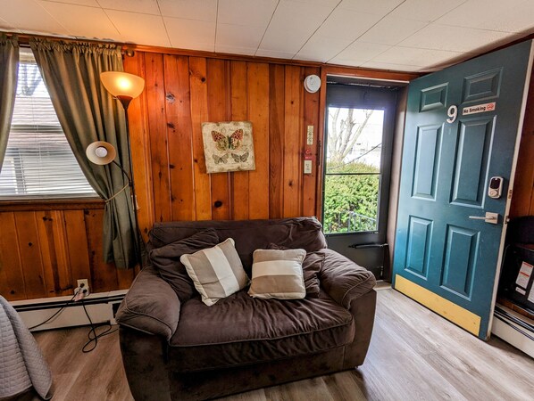 A cozy room with a brown leather sofa, a wooden wall, a framed butterfly artwork, a door with a 'No Smoking' sign, and a window with curtains. - Riverside Motel (Lewiston)
