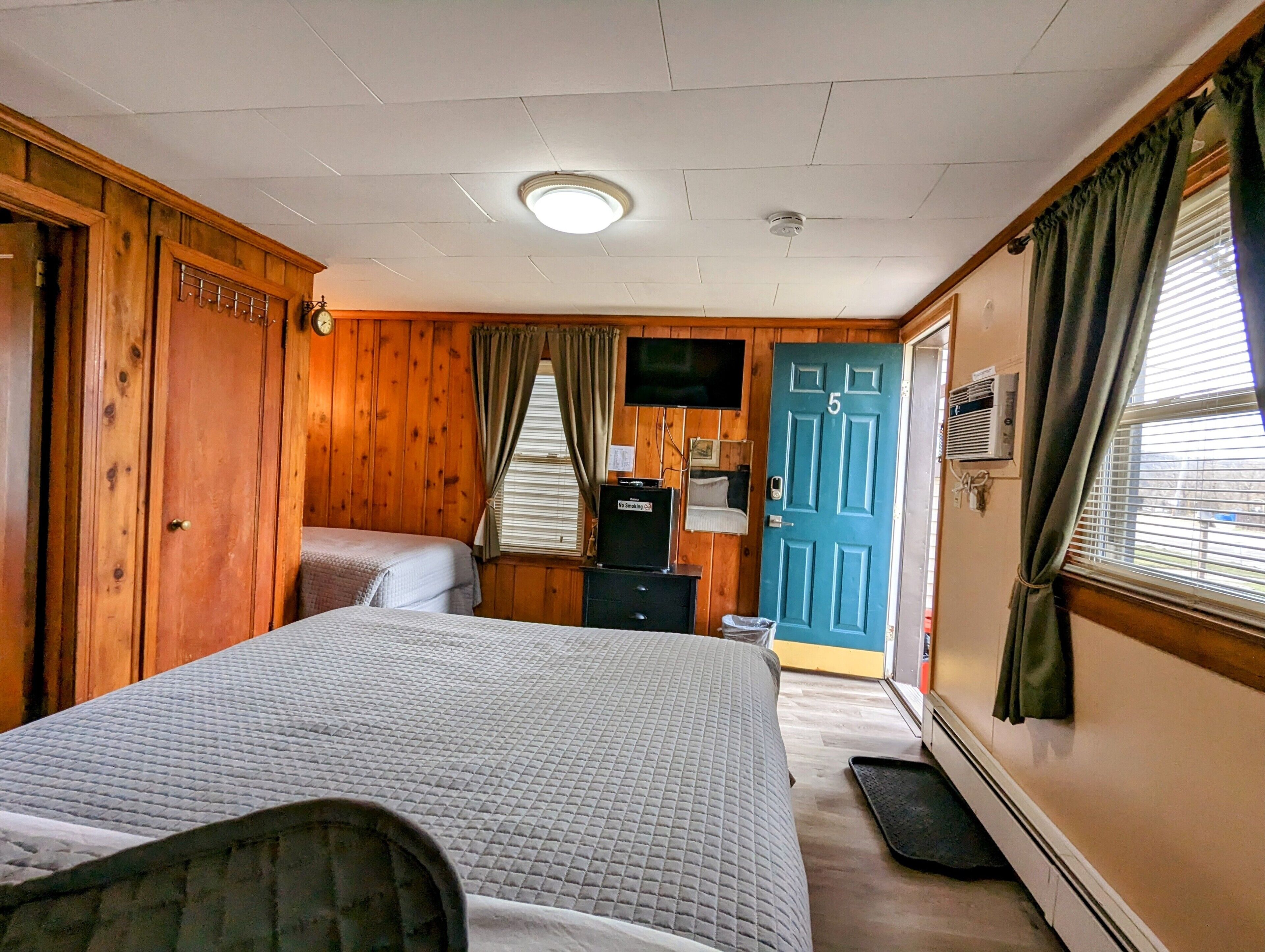 A room with wooden paneling, a bed, a desk, a television, and a door marked with the number 5.