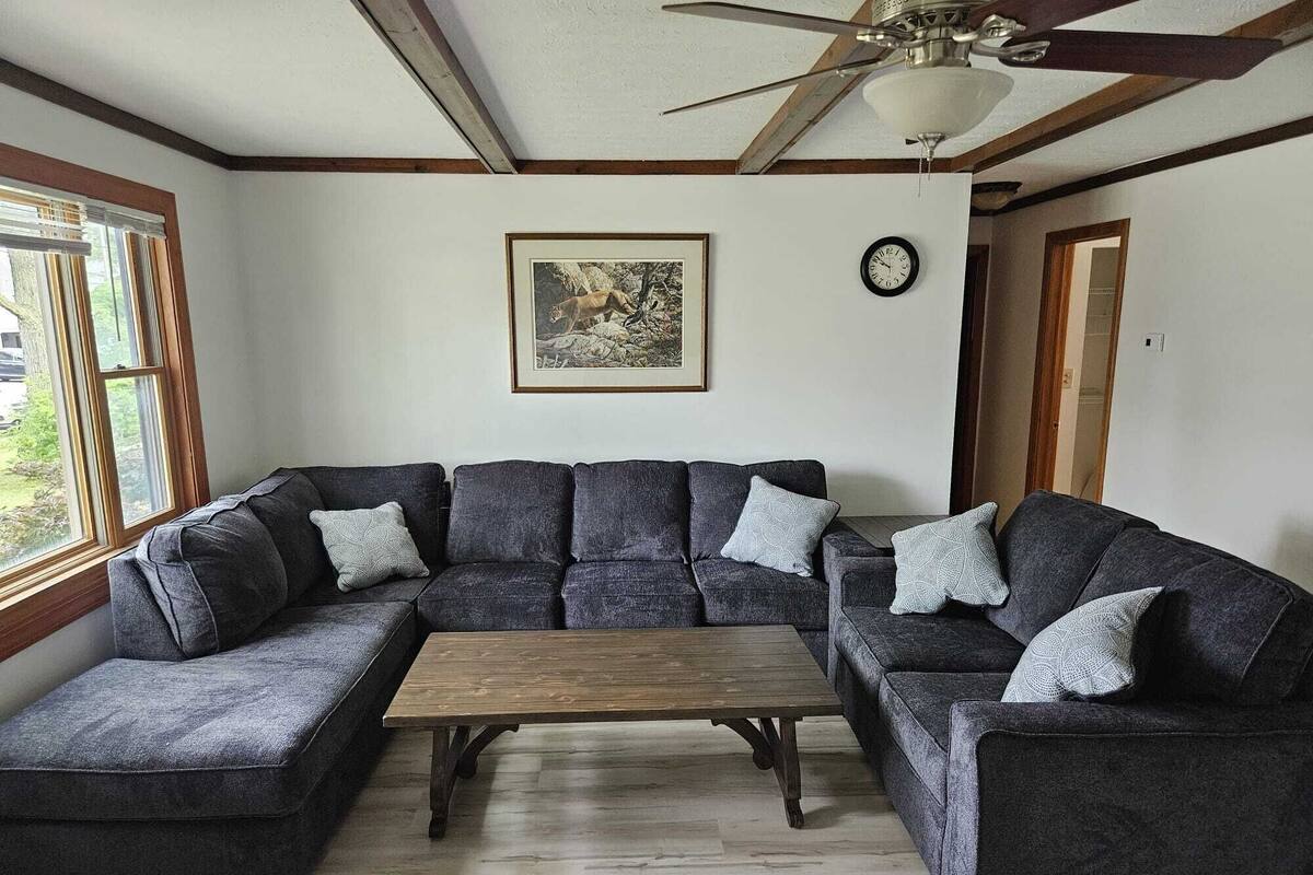 A living room with a sectional sofa, a wooden coffee table, a ceiling fan, a framed picture, and a wall clock.