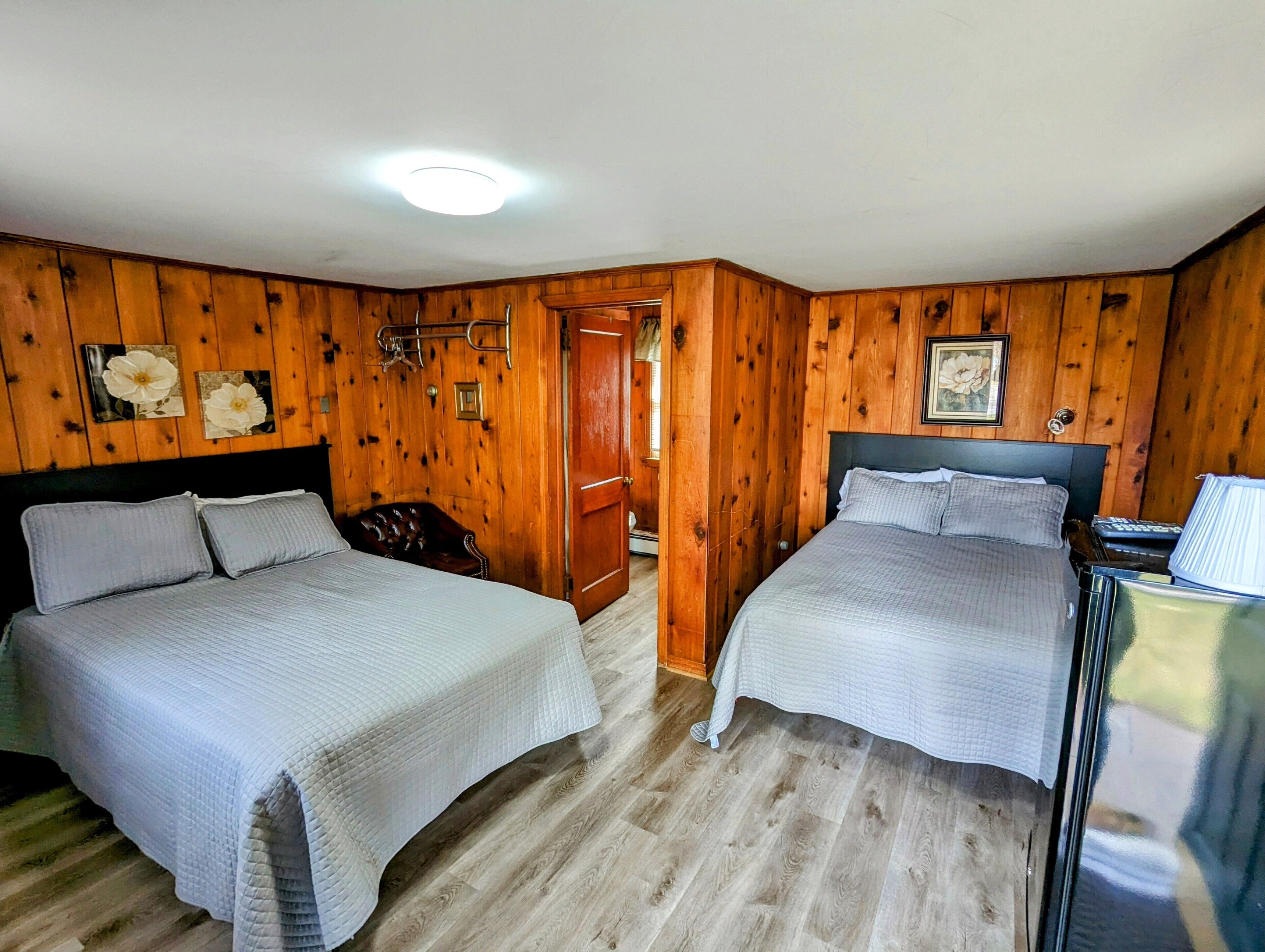 A room with two beds, wooden walls, a ceiling light, and a door leading to another room.