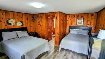 A room with two beds, wooden walls, a ceiling light, and a door leading to another room.