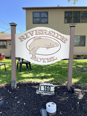 Señal del motel Riverside, con un logotipo de pez, una placa con el número 160 y una casa al fondo.