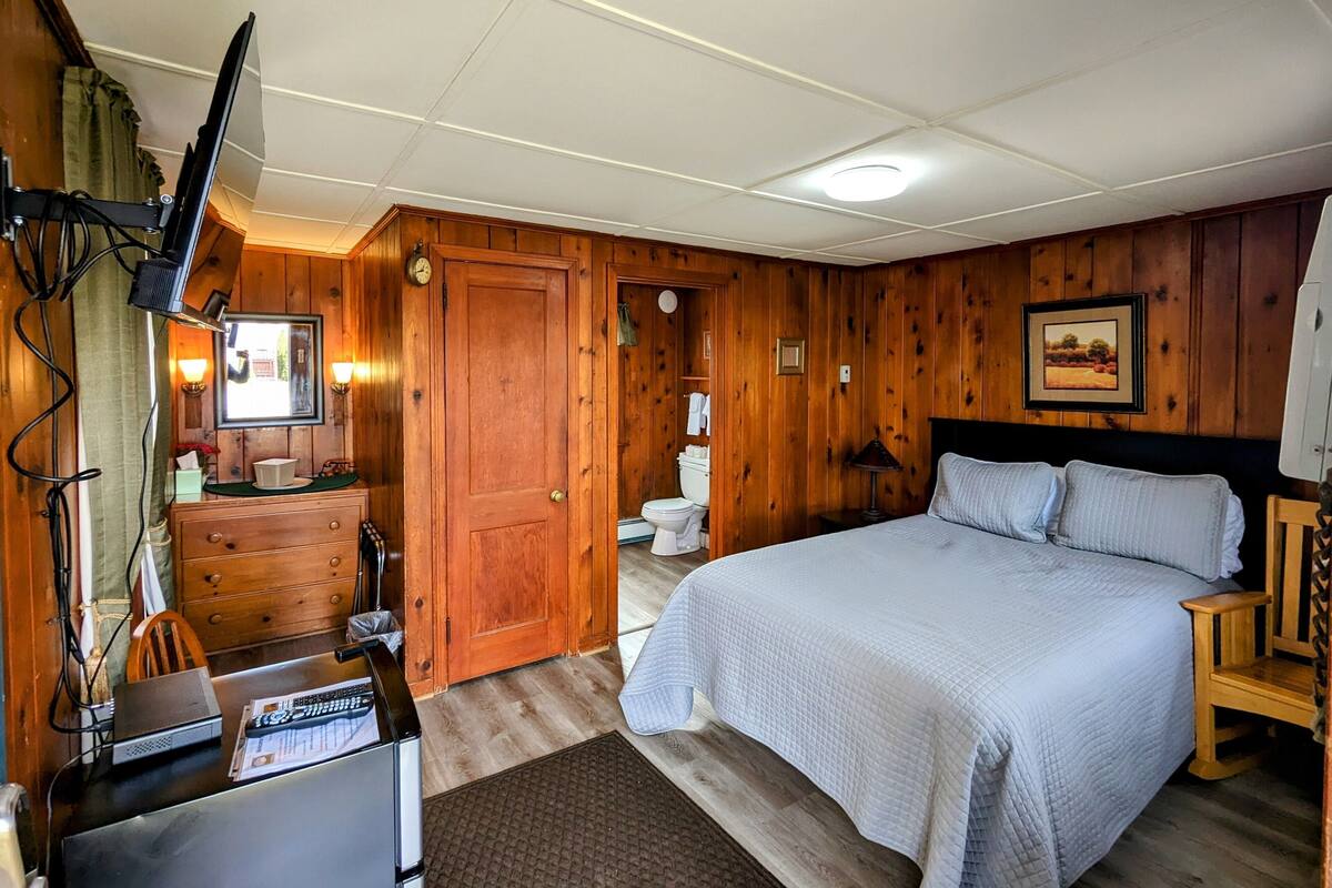 A wooden-paneled room with a bed, a desk with a computer, a television, and a bathroom visible through an open door.