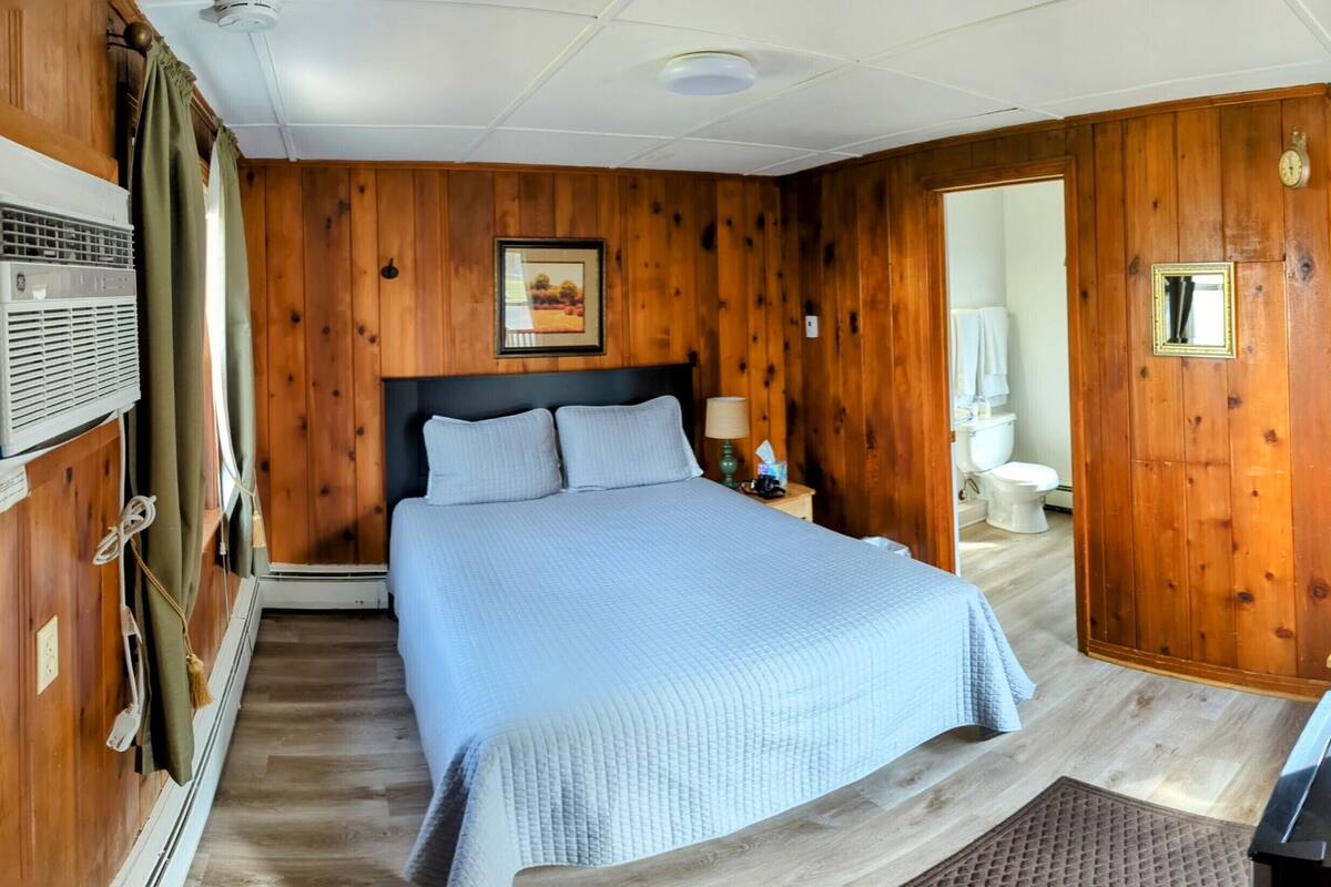 A wooden-paneled room with a bed, a nightstand, a mirror, and a hanging coat rack.