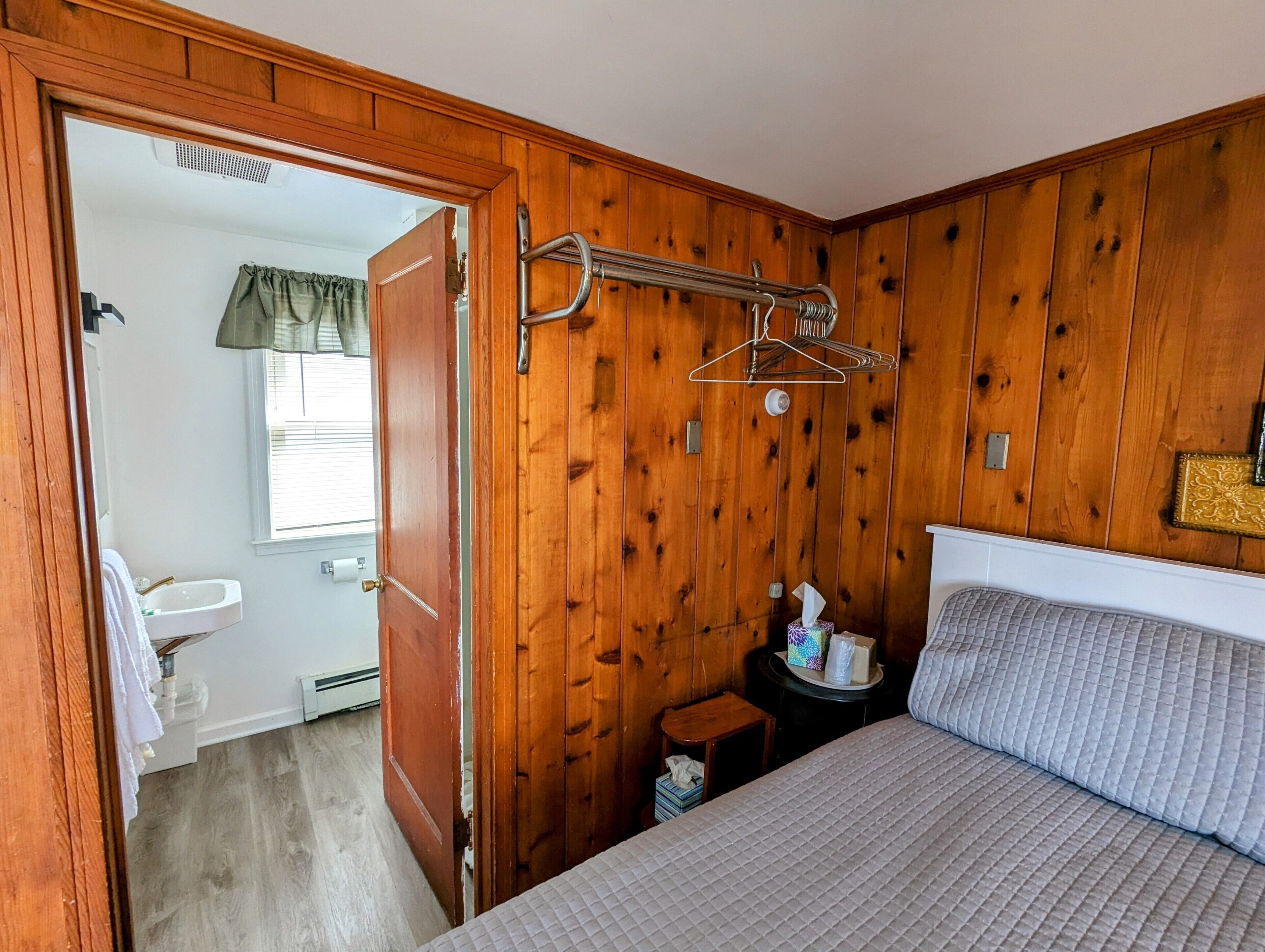 A bedroom with a wooden wall, a bed with a quilted cover, a bedside table with a tissue box, and a hanging clothes rack.