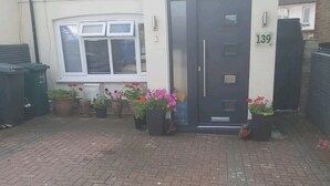 Una casa de dos pisos con una puerta principal oscura, número 139, y un pequeño jardín con plantas y flores en macetas.