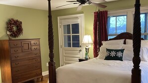 Una cama con dosel con ropa de cama blanca, un aparador de madera con varios cajones, una mesita de noche con una lámpara y una ventana con cortinas rojas.