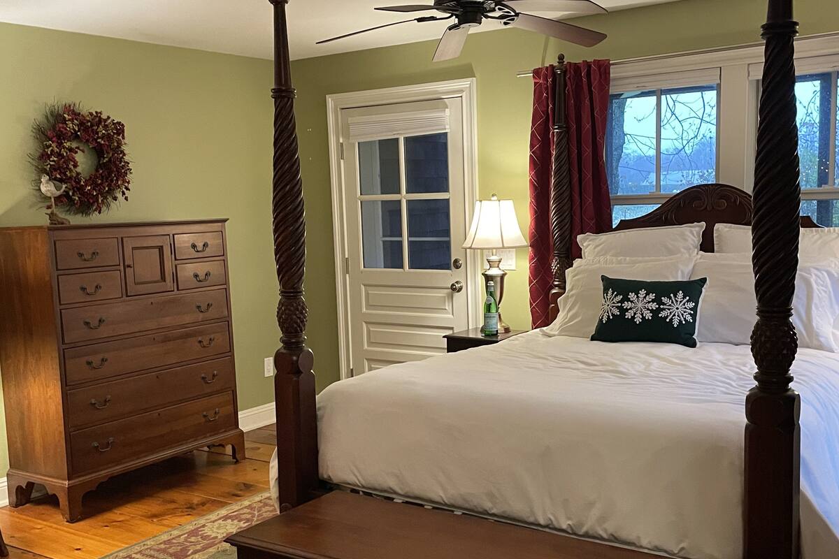 A four-poster bed with white bedding, a wooden dresser with multiple drawers, a nightstand with a lamp, and a window with red curtains.