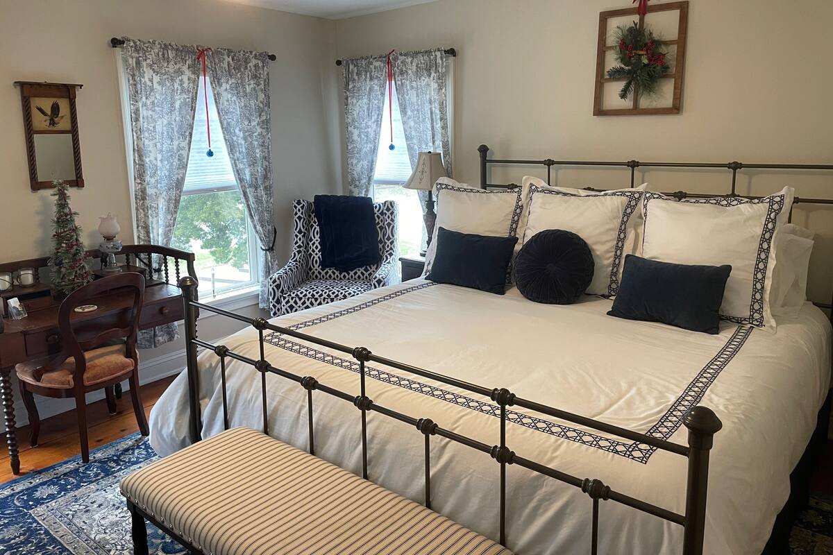 A neatly arranged bedroom with a large bed, a chair, a desk, and a small Christmas tree.