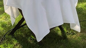 Una mesa redonda con mantel blanco, un expositor de cristal con un pastel blanco, una jarra de cristal, copas de vino y un ramo de flores blancas.