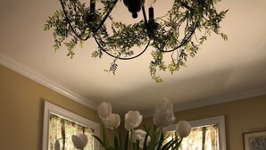 Una mesa de comedor con un florero de flores blancas, rodeada de sillas, en una habitación con ventanas y una araña.