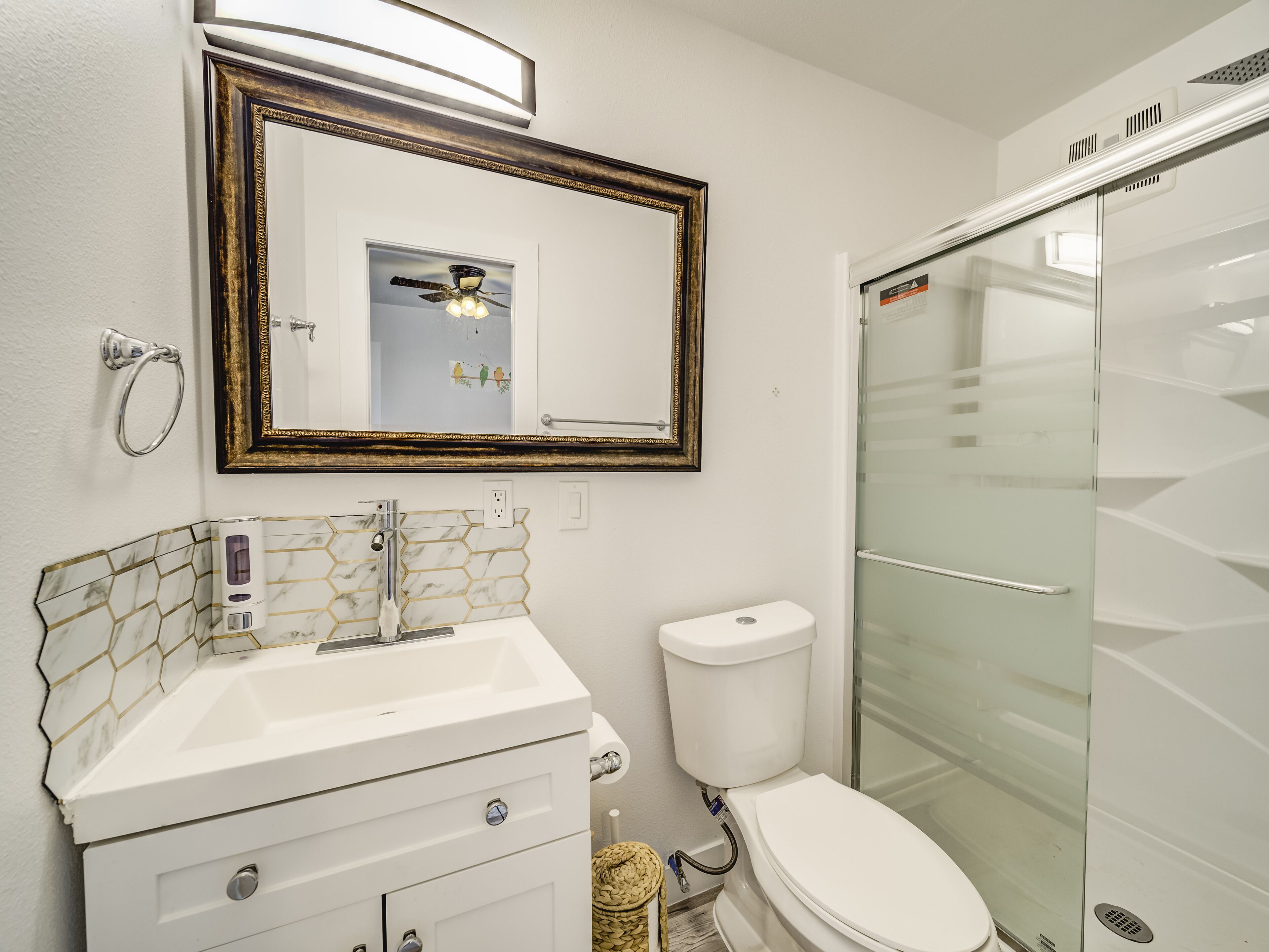 Une salle de bain avec un lavabo blanc, des toilettes, une cabine de douche en verre et un miroir doté d’un cadre décoratif.