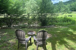 To Adirondack-stole og et lille bord i et græsklædt udendørs område med træer og udsigt over et mark.