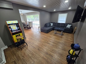 A living room with a sectional sofa, a coffee table, a Pac-Man arcade game, and a TV mounted on the wall.
