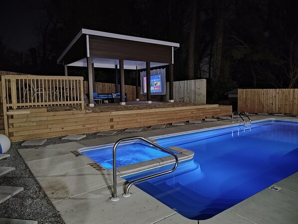 A swimming pool with a covered patio area, wooden deck, and a wooden fence.