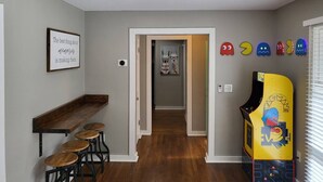 Un pasillo con un banco y taburetes de madera, un ventilador de techo y una máquina arcade Pac-Man.