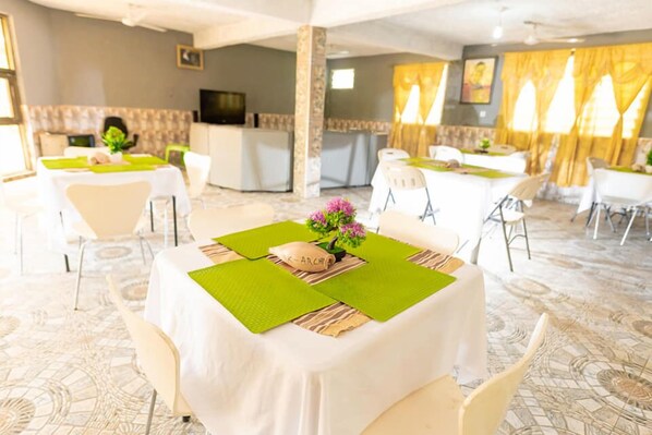 A hotel dining area with tables set for guests, a kitchen in the background, and a patterned carpet. - K-archy Lodge (Kyebi)