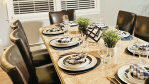 Une salle à manger avec une longue table dressée pour six personnes, agrémentée d’un lustre moderne, d’une grande fenêtre avec stores et d’un tableau représentant un paysage urbain, encadré et accroché au mur.
