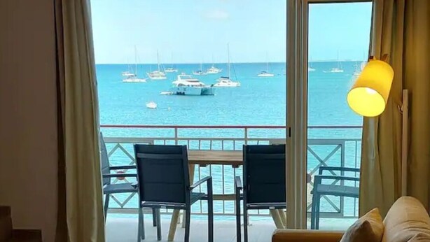 Een balkon met uitzicht op zee en boten, een bank, een tafel met een kom en een lamp.